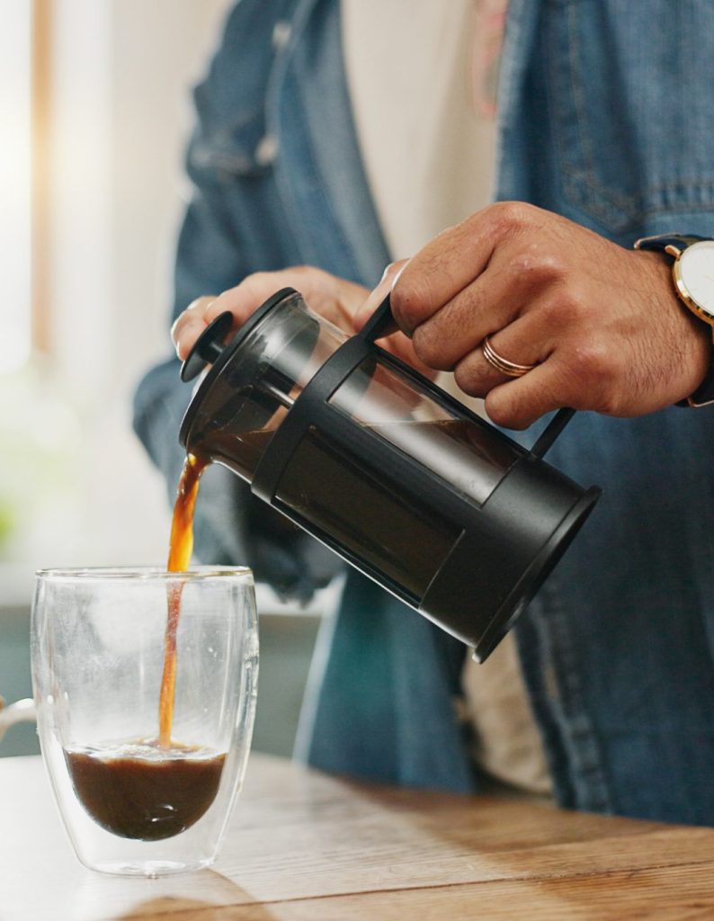 Hands, pouring coffee and breakfast in home kitchen, energy and start morning with warm matcha at table. Man, filter kettle and french press plunger for fresh organic caffeine at house for hot drink.