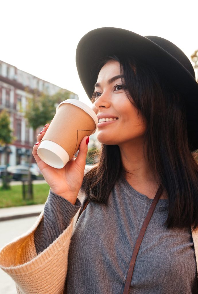 Smiling stylish young woman drinking coffee while walking on a city street